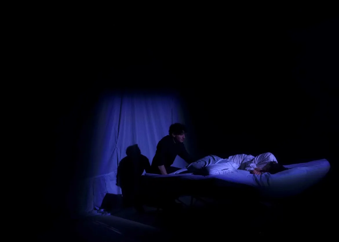The Duchess kneeling forward on her bed in blue light.