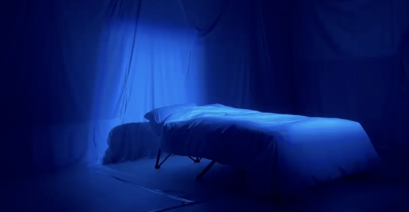 Empty bed lit blue, drapes falling.