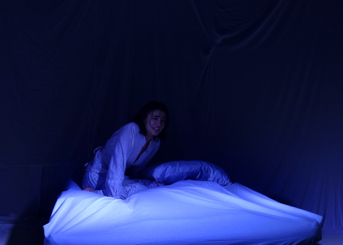 The Duchess kneeling on her bed in a wash of cold blue light.