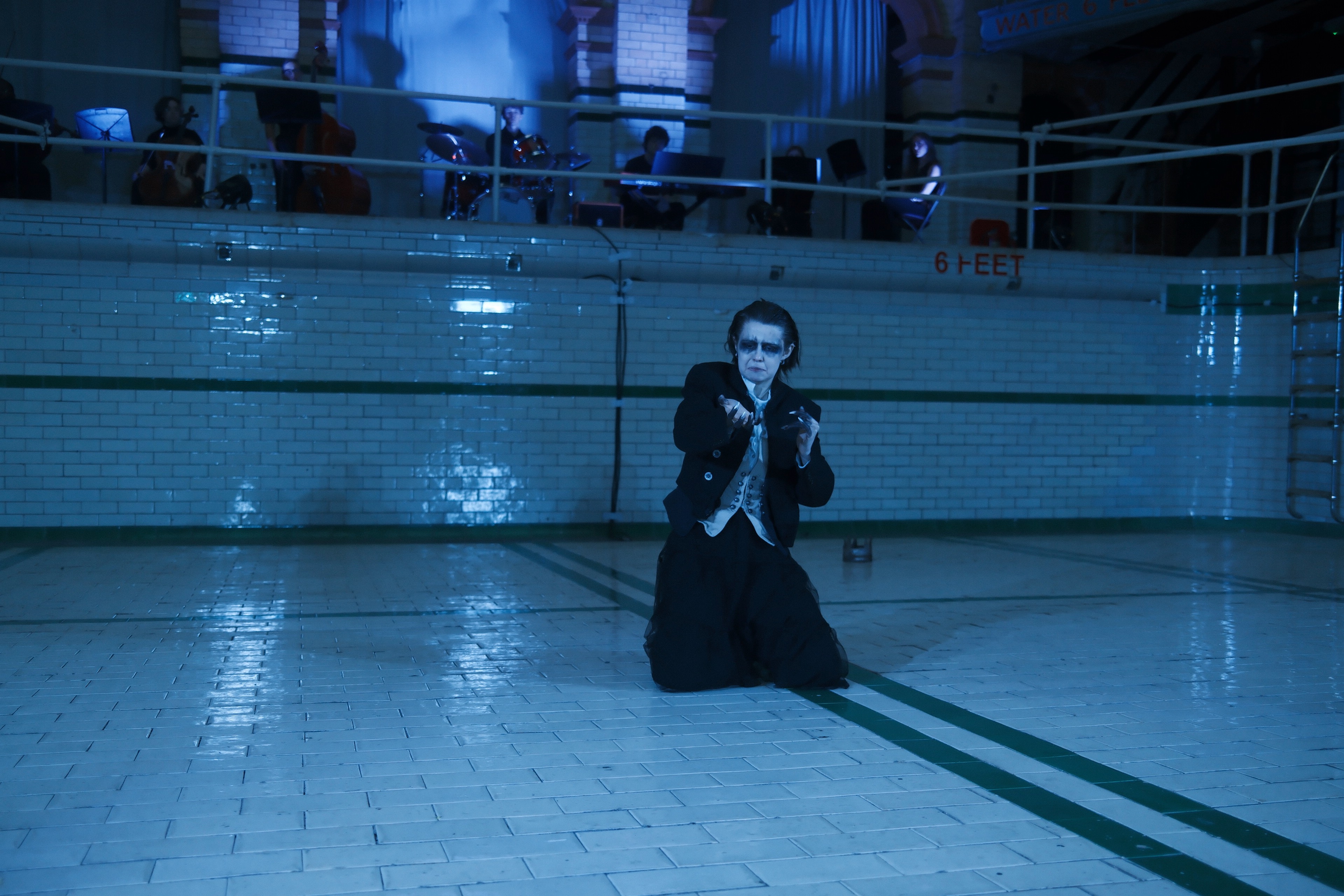 Richard III kneeling alone under cold blue wash, band behind on the balcony.