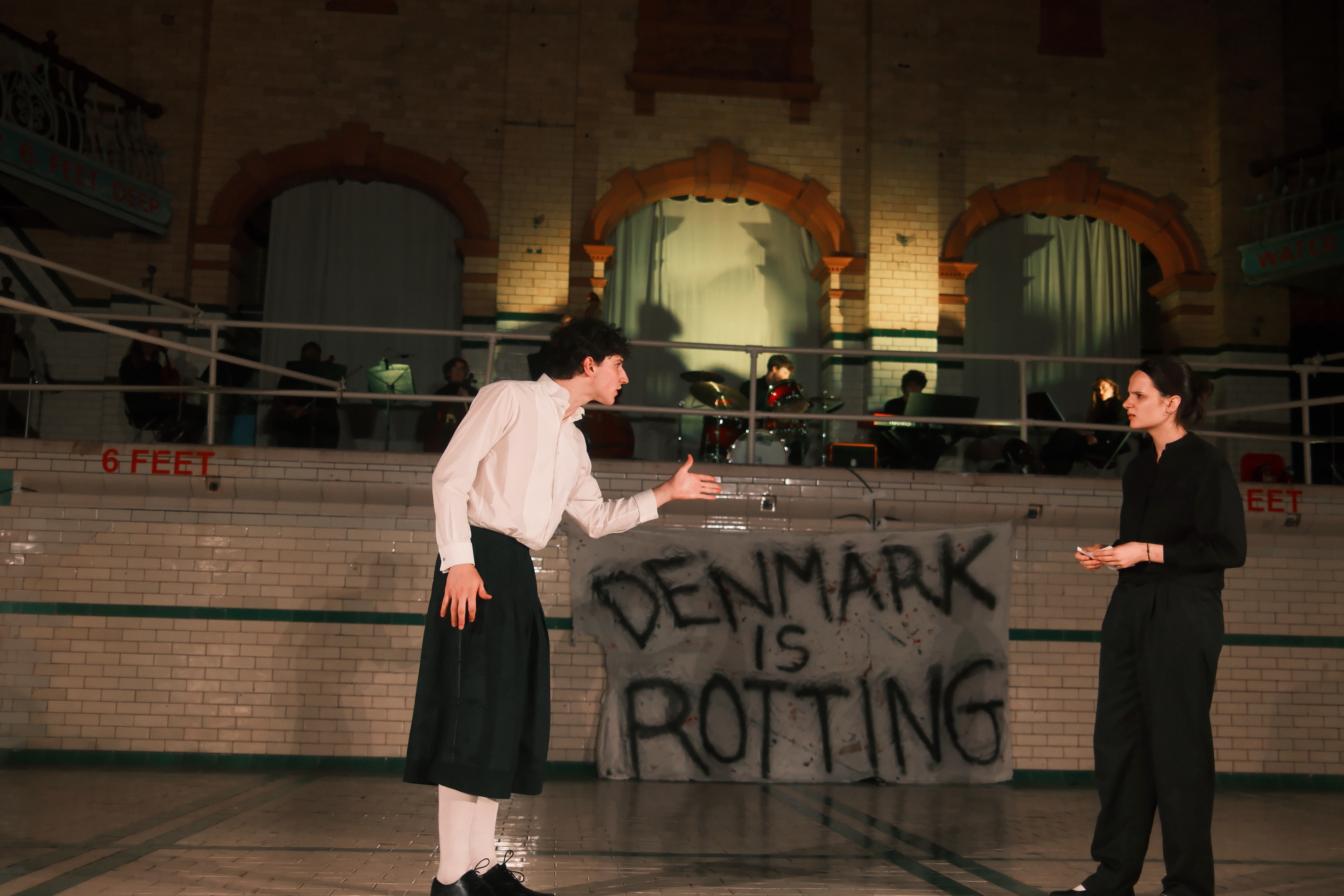 Hamlet gesturing to Gertrude in front of a hand-painted 'Denmark Is Rotting' banner.
