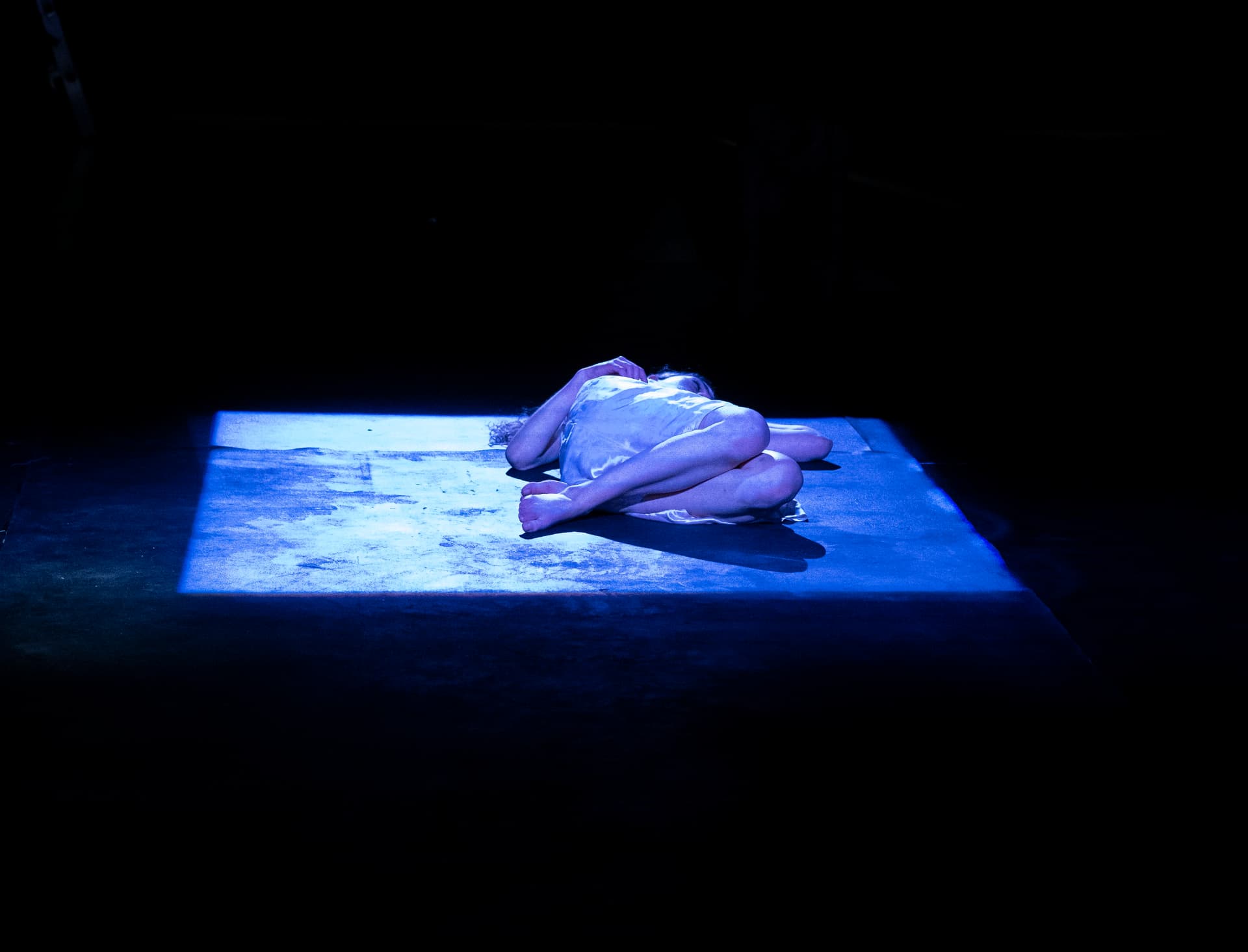 Eurydice curled in a lit square on a black stage.